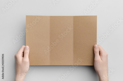 Kraft A4 Tri-Fold Brochure Mock-Up - Male hands holding a kraft tri-fold on a gray background. © Shablon