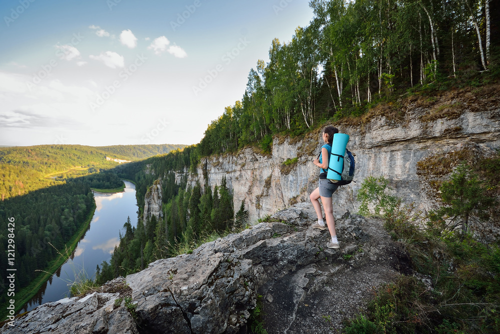 Young woman hiker with backpack standing on cliff and looking far away enjoying beautiful view