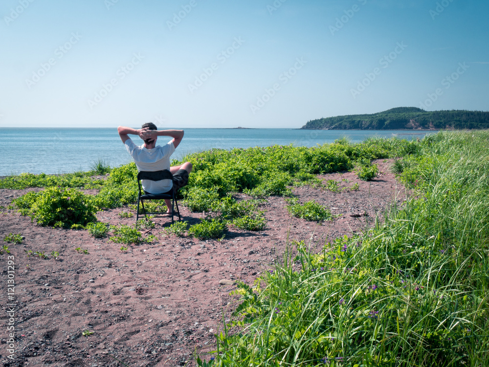Fototapeta premium Man sitting on chair on stony beach overlooking a bay with hills