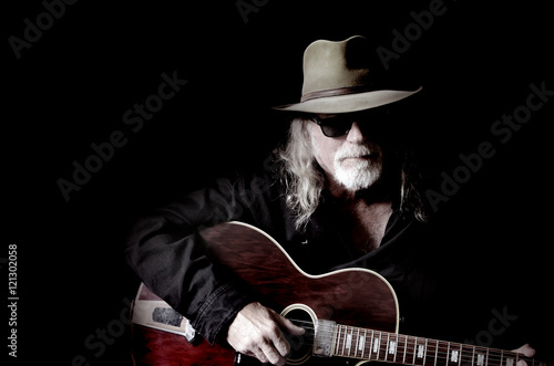 Middle-aged Caucasian man with long hair and a white beard wearing sunglasses, a hat and black shirt while holding a guitar. Black background and sidelighting. Vintage filter and desaturation applied