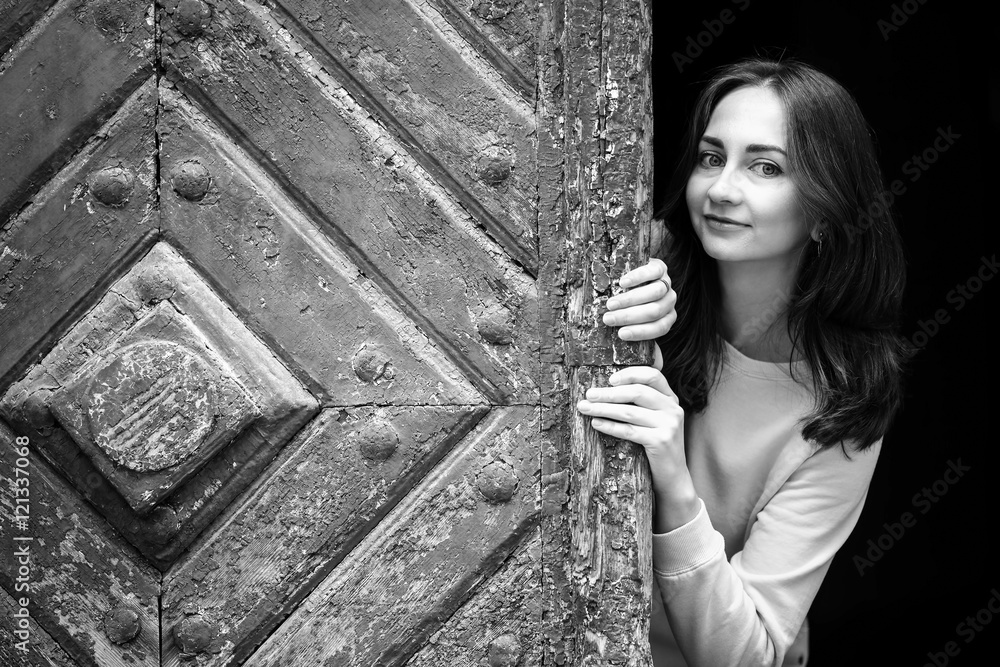 Obraz premium Pretty young girl peering from behind ancient wooden door, black-and-white photo.