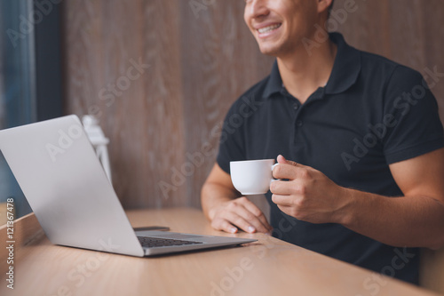 Casual man working on laptop and holding cup of coffee while sitting in cafe