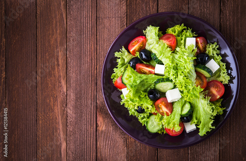Greek salad (lettuce, tomatoes, feta cheese, cucumbers, black olives) on dark wooden background top view. Space for text. Healthy food.