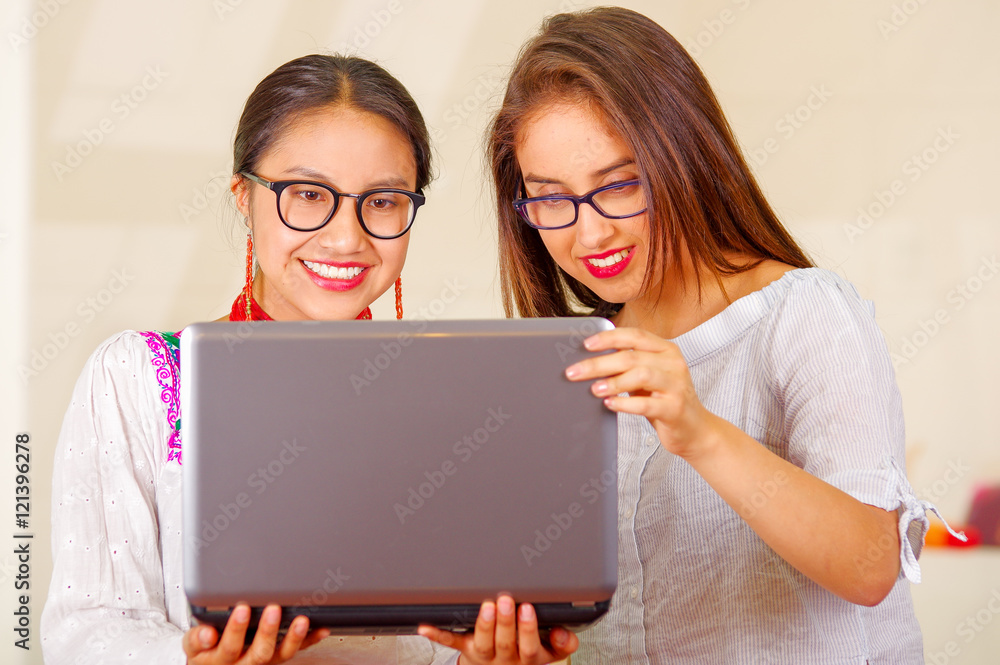 Two beautiful young women posing for camera, one wearing traditional andean clothing, the other in casual clothes, holding laptop between them interacting looking at screen, both smiling, park