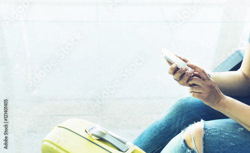 Young hipster girl sitting at airport in yellow boot on suitcase traveling, female hands using app on making smartphone in terminal area hall, tourist journey trip, mockup blank screen sell phone