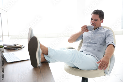 Relaxed handsome man is drinking coffee having put his legs on table