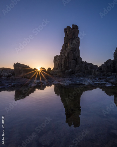 Sun flare as the sun rises behind a reflection of rocks in a rock pool at Bombo Quarry, New South Wales, Australia
