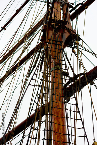 The wooden mast and lots of ropes on a large sailboat. Gray sky above.