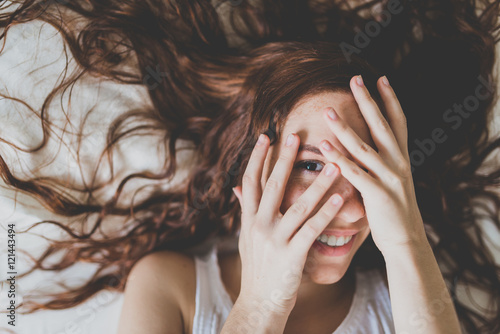 Red-haired woman covering face with hands and looking at camera with toothy smile photo