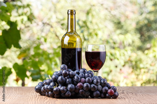 Bottle and a glass of wine with grapes on a wooden background outdoors.