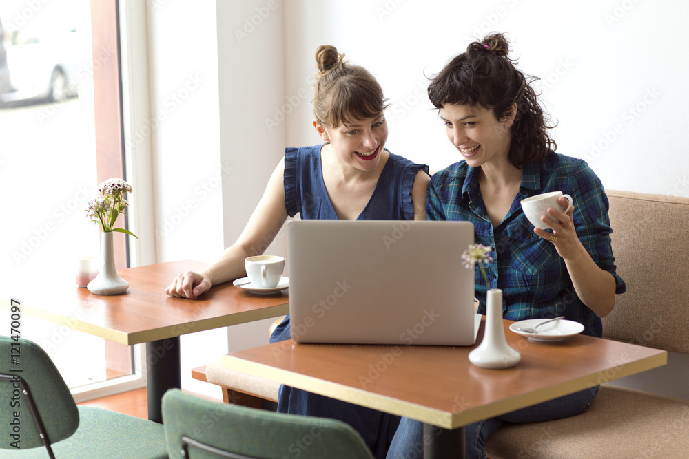 Two smiling friends sitting in a coffee shop looking at laptop Two smiling friends sitting in a coffee shop looking at laptop