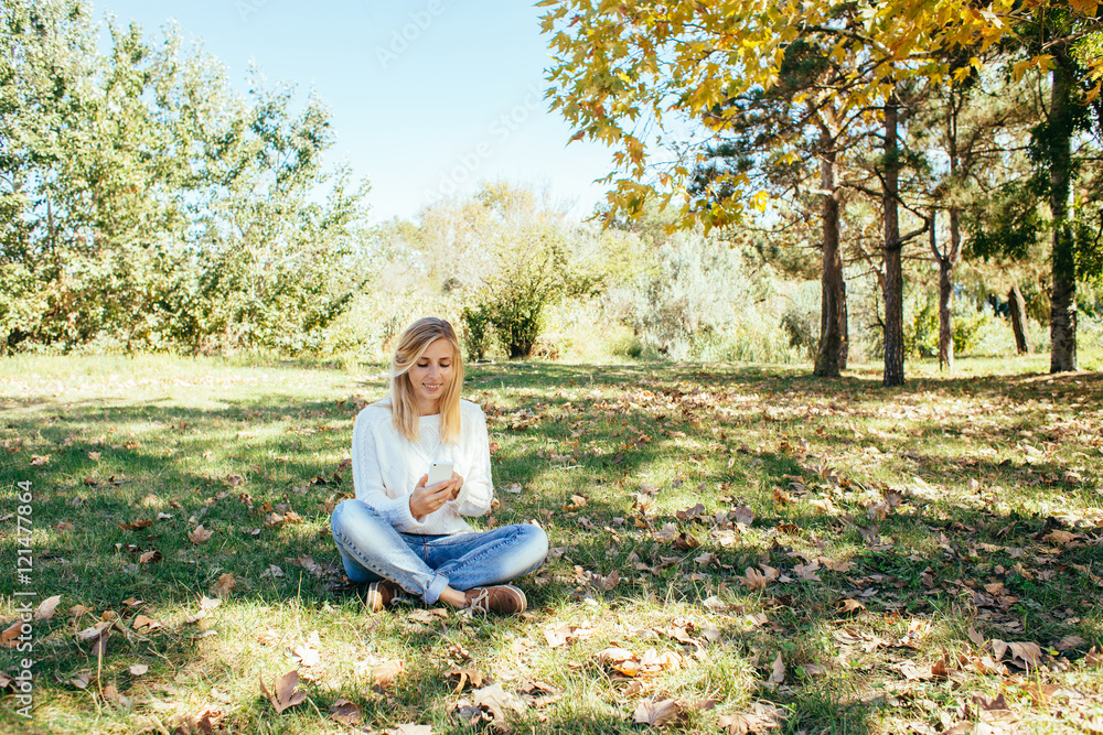 young woman in a white sweater and blue jeans sitting in the park and using her cell phone. woman sending a text message from her mobile phone outdoor young woman in a white sweater and blue jeans sitting in the park and using her cell phone. woman sending a text message from her mobile phone outdoor