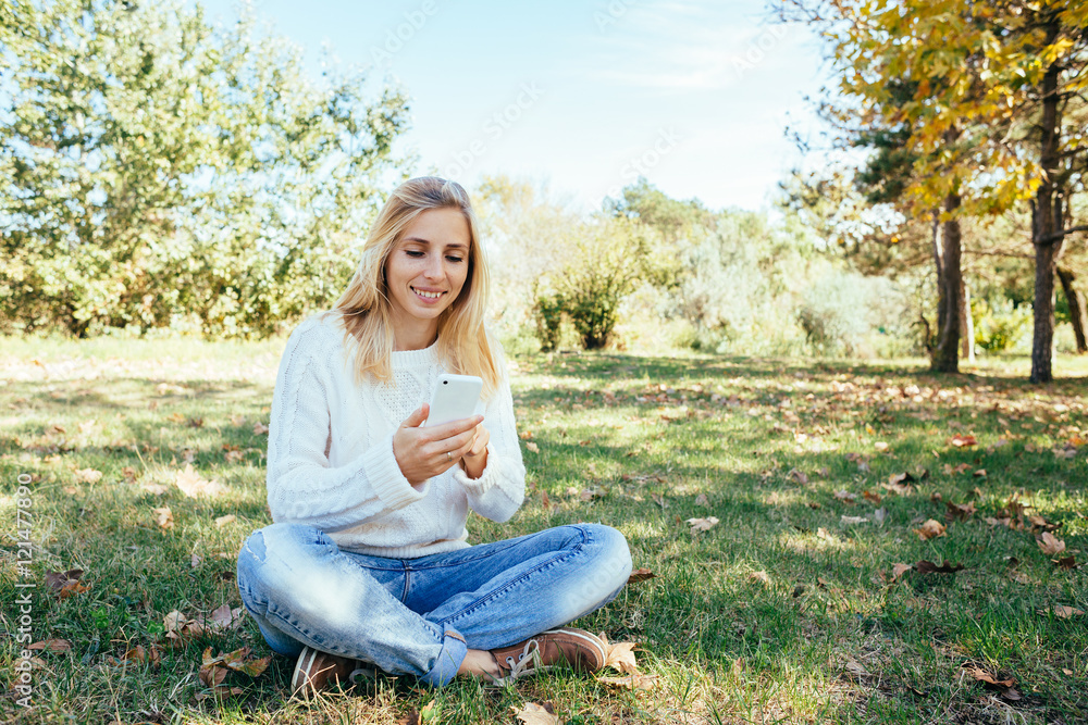young woman in a white sweater and blue jeans sitting in the park and using her cell phone. woman sending a text message from her mobile phone outdoor young woman in a white sweater and blue jeans sitting in the park and using her cell phone. woman sending a text message from her mobile phone outdoor