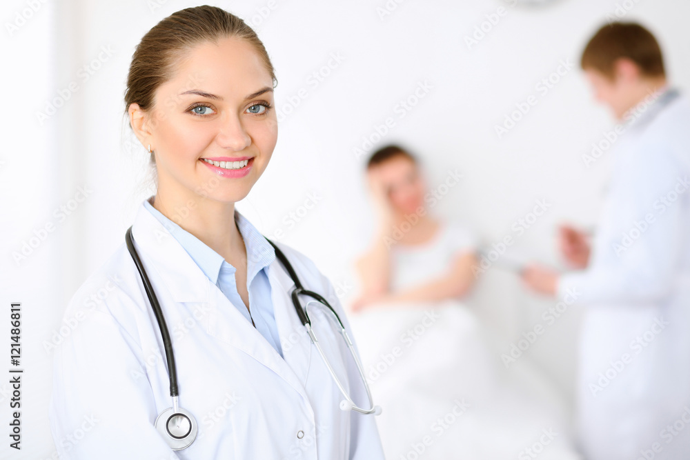 Cheerful smiling female doctor on the background with physician and his patient in the bed. High level and quality of medical service concept. Cheerful smiling female doctor on the background with physician and his patient in the bed. High level and quality of medical service concept.