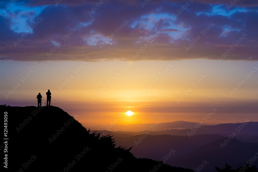 Silhouettes of two people standing on a rock and looking toward the sun. Dramatic clouds over them. Sunset in the mountains. Purple light. Summer in the Ukrainian Carpathians Silhouettes of two people standing on a rock and looking toward the sun. Dramatic clouds over them. Sunset in the mountains. Purple light. Summer in the Ukrainian Carpathians