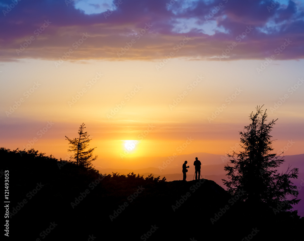 Silhouettes of two people standing on a rock near the tree and looking toward the sun. Sunset in the mountains. Summer in the Ukrainian Carpathians