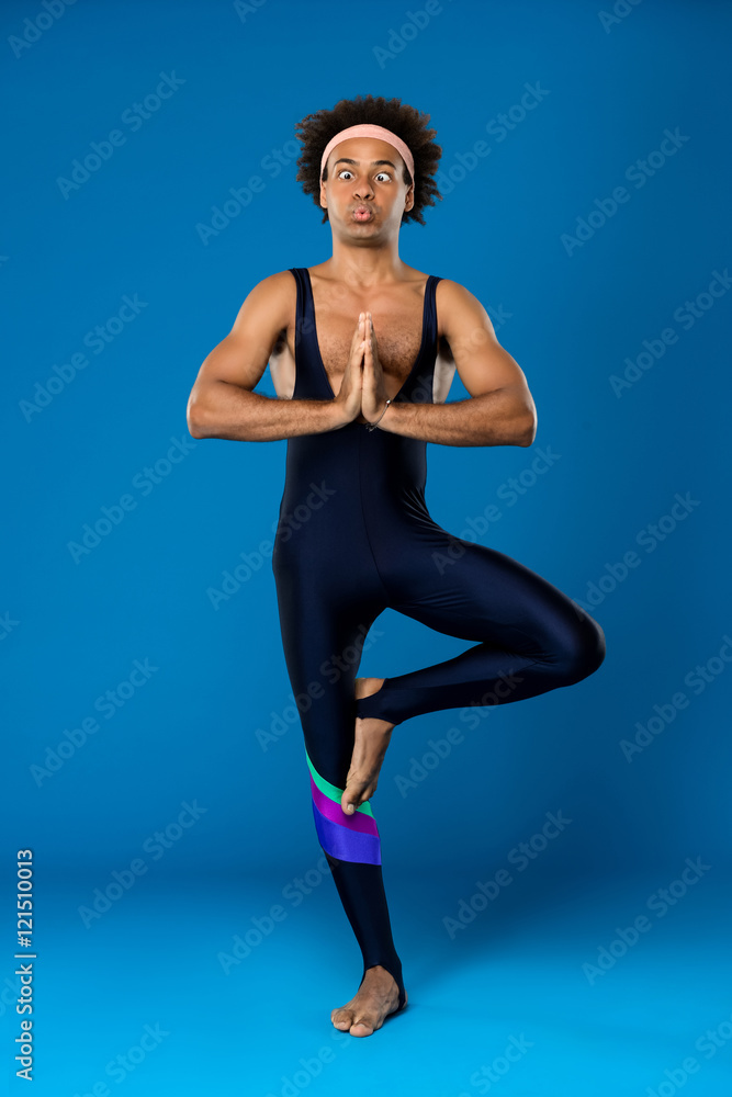 Sportive african man training yoga Vrikshasana, fooling over blue background.