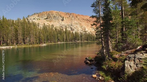 Aerial shot flying over a mountain lake in the Sierra Nevada mountains. Green trees and turquoise blue-green water with a mountain overlooking. Fallen trees can be seen under the water. 