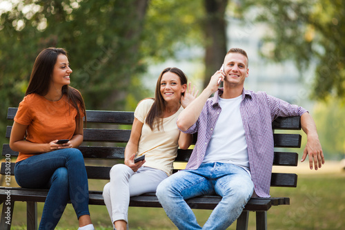 Three friends are sitting on bench in park and using smartphones.
