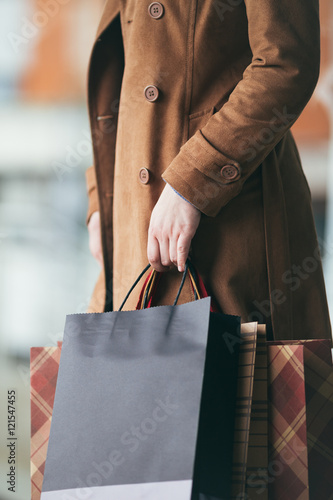 Elegant and modern woman in brown leather jacket or topcoat standing on the city street and holding bunch of shopping bags. Close up shot of woman hand. Lifestyle consumerism theme. 