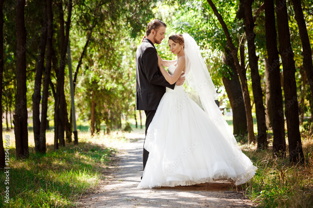 Young beautiful dressy newlyweds smiling, posing, embracing in park.