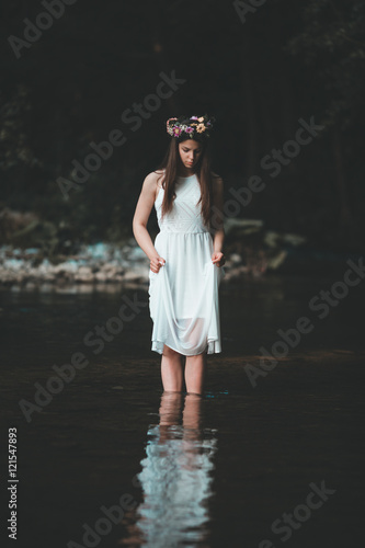 Outdoor portrait of beautiful young woman wearing white dress and floral wreath on her head. Girl walking alone through cold mountain river.