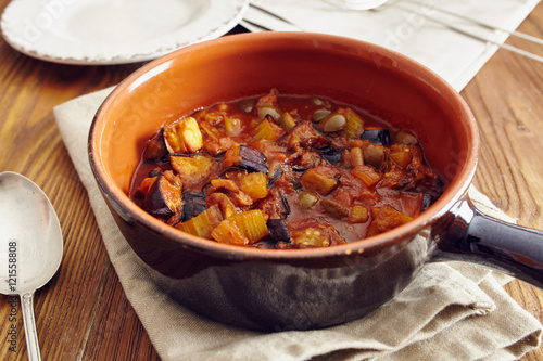 eggplant caponata, traditional sicilian dish. Served in a classic ceramic pan over a napkin on an aged wooden table. Surrounded by a silver spoon and a dish photo