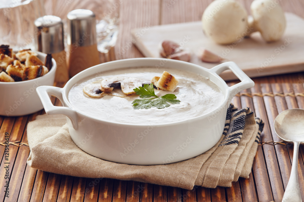Mushroom cream soup on a table, with bread, garlic, and raw mushrooms on a white chopping board. Salt and Pepper on the table..