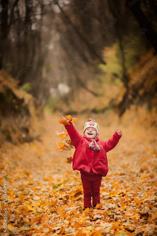 Little girl in red coat at scenic fall park. Baby wearing knitted cap standing over foliage background. Toddler child playing with yellow autumn leaves. Portrait of cute funny child outside. Stock Pho...