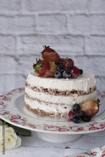 Naked cake with caramelized fruits - strawberries, blueberries, raspberries. Sponge cream cake in floral high plateau, tray. White roses, rustic background
