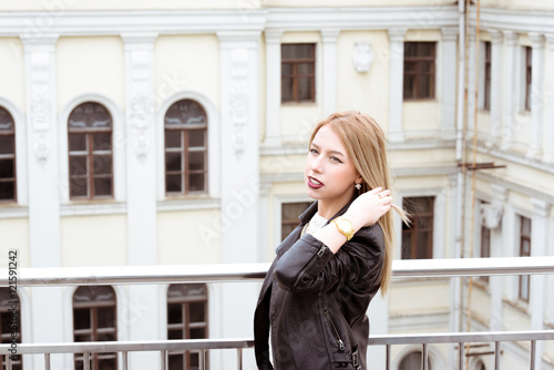Beautiful blond young hipster girl with red lipstick and bright makeup on a sunny day outdoors. Smiling posh sexy woman dressed in a black leather jacket posing near old buildings 