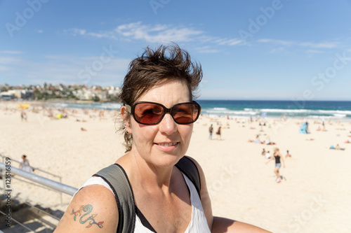 A middle aged woman in sunglasses enjoying visiting Bondi Beach in Sydney, Australia, with short hair, sunglasses, and a tattoo on her shouler.
