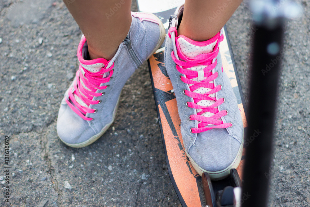 Accurate close-up view athlete young woman on a kick scooter is placed her foots in grey shoes with pink laces on bike on a background of asphalt Accurate close-up view athlete young woman on a kick scooter is placed her foots in grey shoes with pink laces on bike on a background of asphalt