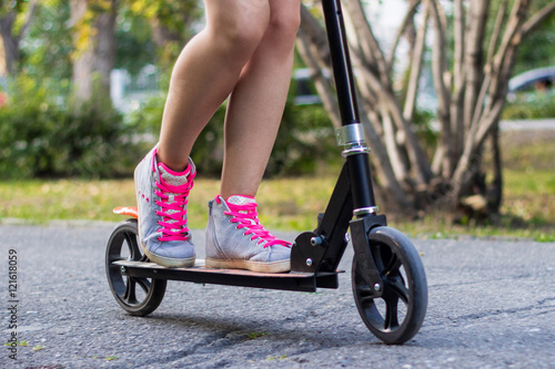 Athlete female teenager in gray shoes with pink laces is riding on a black kick scooter on asphalt road in a park with landscape on a background. Close-up horizontal view