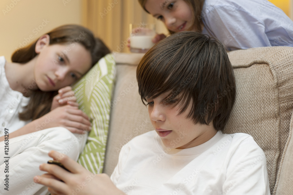 Teenage boy using smartphone while sisters watch in background Teenage boy using smartphone while sisters watch in background
