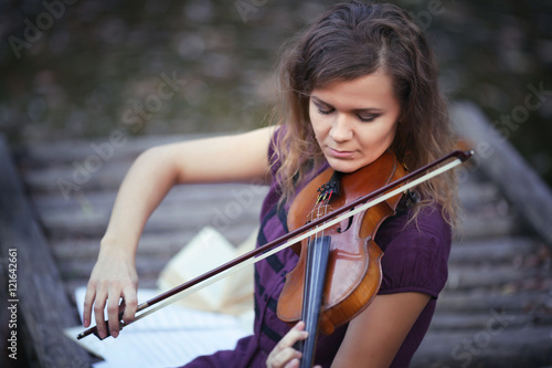 Portrait of young woman playing on the violin outdoors, at lake pier autumn. Around leaves on water. Girl in violet dress musical notes on pier.
