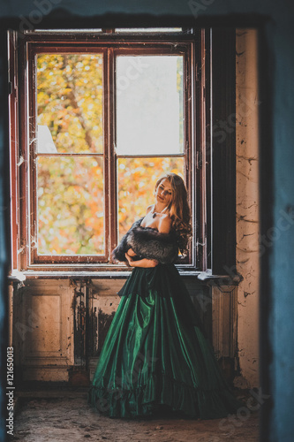 Beautiful girl in green vintage dress with coat walking near ancient house and door. Vintage farytale concept. Woman with long hair and make up