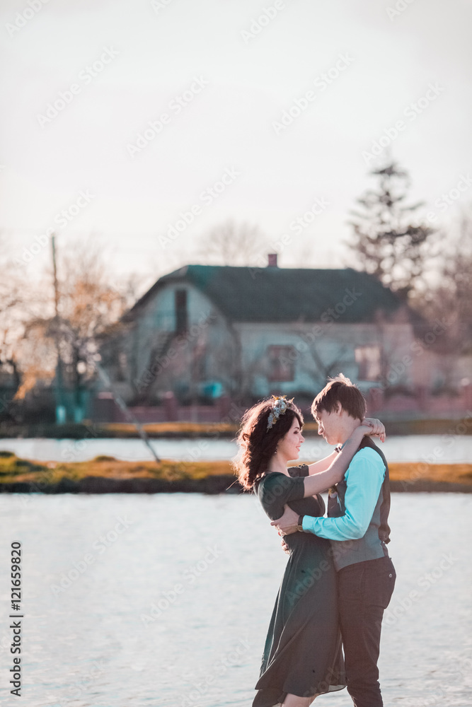Charming bride, elegant groom on landscapes of mountains and sunset at lake. Gorgeous wedding couple. Man in blue shirt and waistcoat. Woman in  green dress and crown.