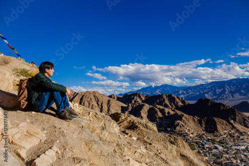 Young Asian traveler sitting on high hills and looking away at the village over blue sky background in Leh, Ladakh, India