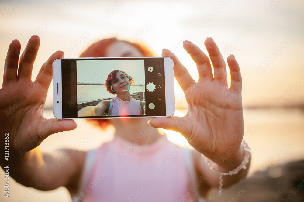 girl in pale pink dress with red hair and backpack walking along river bank, pictures of themselves on their mobile camera phone, against backdrop of boats moored on a warm summer day girl in pale pink dress with red hair and backpack walking along river bank, pictures of themselves on their mobile camera phone, against backdrop of boats moored on a warm summer day