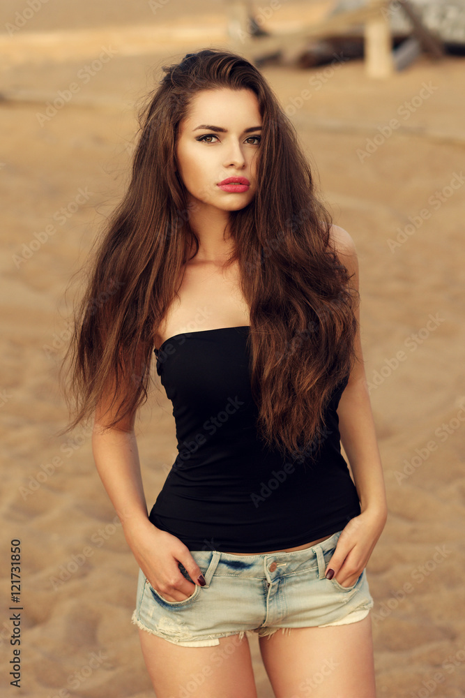 Outdoor portrait of young stylish woman wearing black t-shirt or top and  blue jeans shorts standing on the beach. Pretty sexy hipster girl Stock  Photo