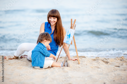 Fotografía Woman and child in blue and white dress sit on the beach and draw on the easel
