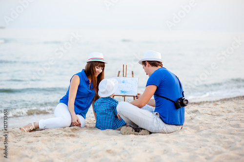 Woman, man and child in blue and white dress sit on the beach and draw on the easel near the surf © freshstockplace