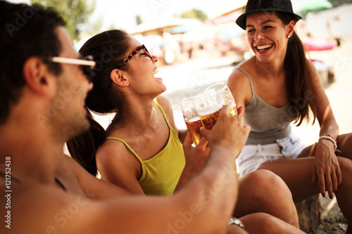 Group of young friends sitting on  sunbeds at the riverside beach, relaxing and drinking beer.Summer beach party. © BalanceFormCreative