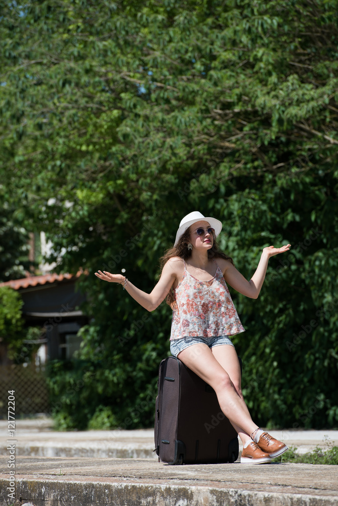 Naklejka premium Pretty girl with curly hair wear casual clothes, sunglasses and panama hat, waiting with rolling upright suitcase at old docks train station