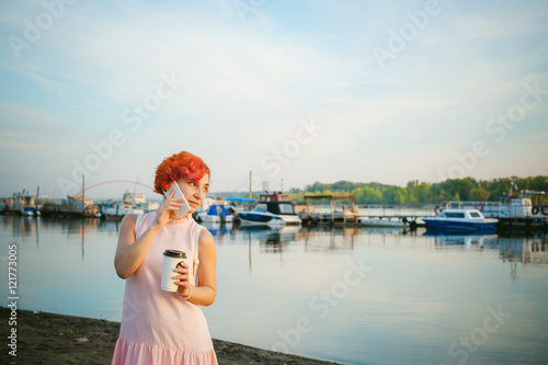 girl in pale pink dress with red hair and backpack walking along river bank, talking on the phone and drinking coffee from a cardboard cup, against backdrop of boats moored on a warm summer day © evgeniykleymenov