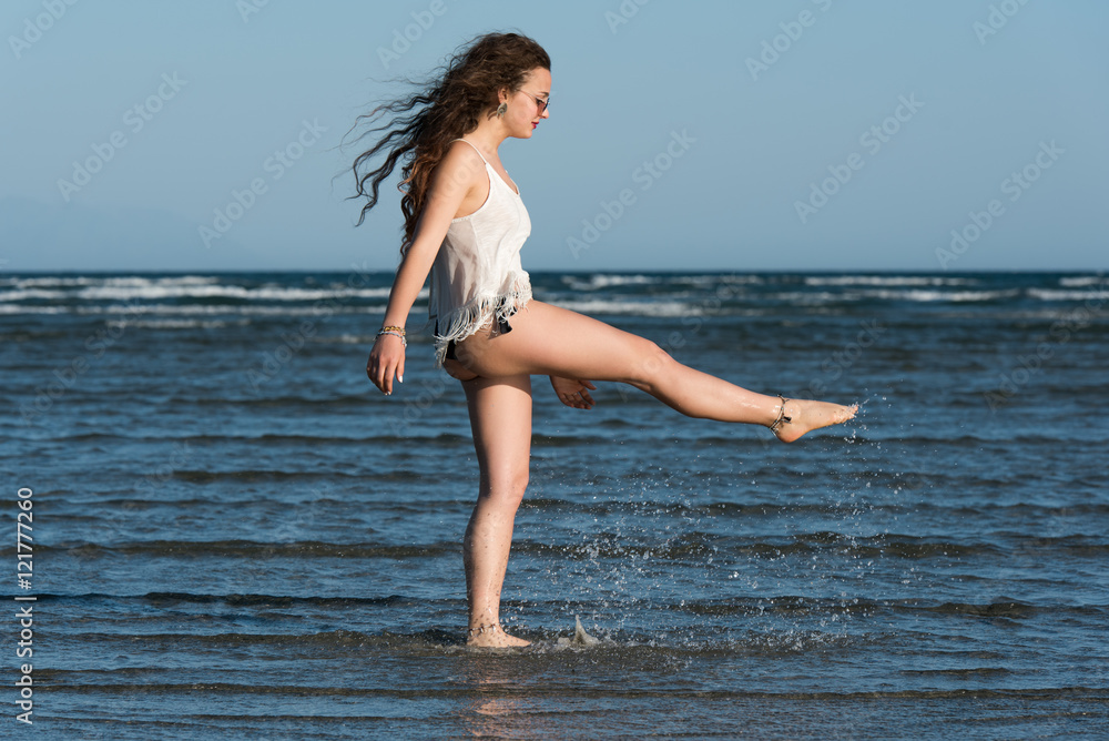 Naklejka premium Woman with long curly hair wear bikini and shirt, standing in sea and kick the water. Sea and sky as background