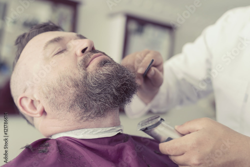 Male barber cuts the beard using clipper of a adult man with a mohawk. Toned