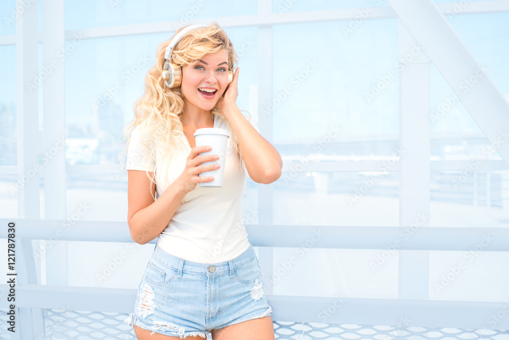 Morning walks / Beautiful young woman with music headphones, standing on the bridge with a take away coffee cup and posing against urban background. Morning walks / Beautiful young woman with music headphones, standing on the bridge with a take away coffee cup and posing against urban background.