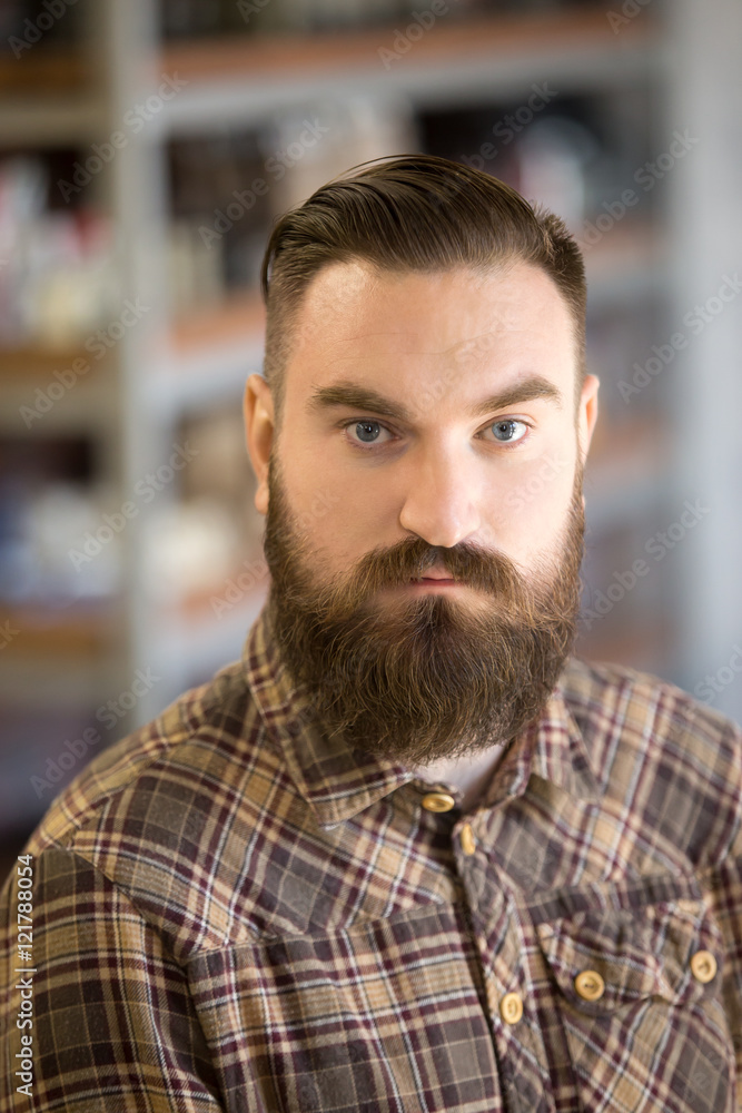 Naklejka premium Headshot portrait of serious young man with long beard and mustache. Handsome bearded man posing with hands crossed on his chest, looking at camera, standing in his shop